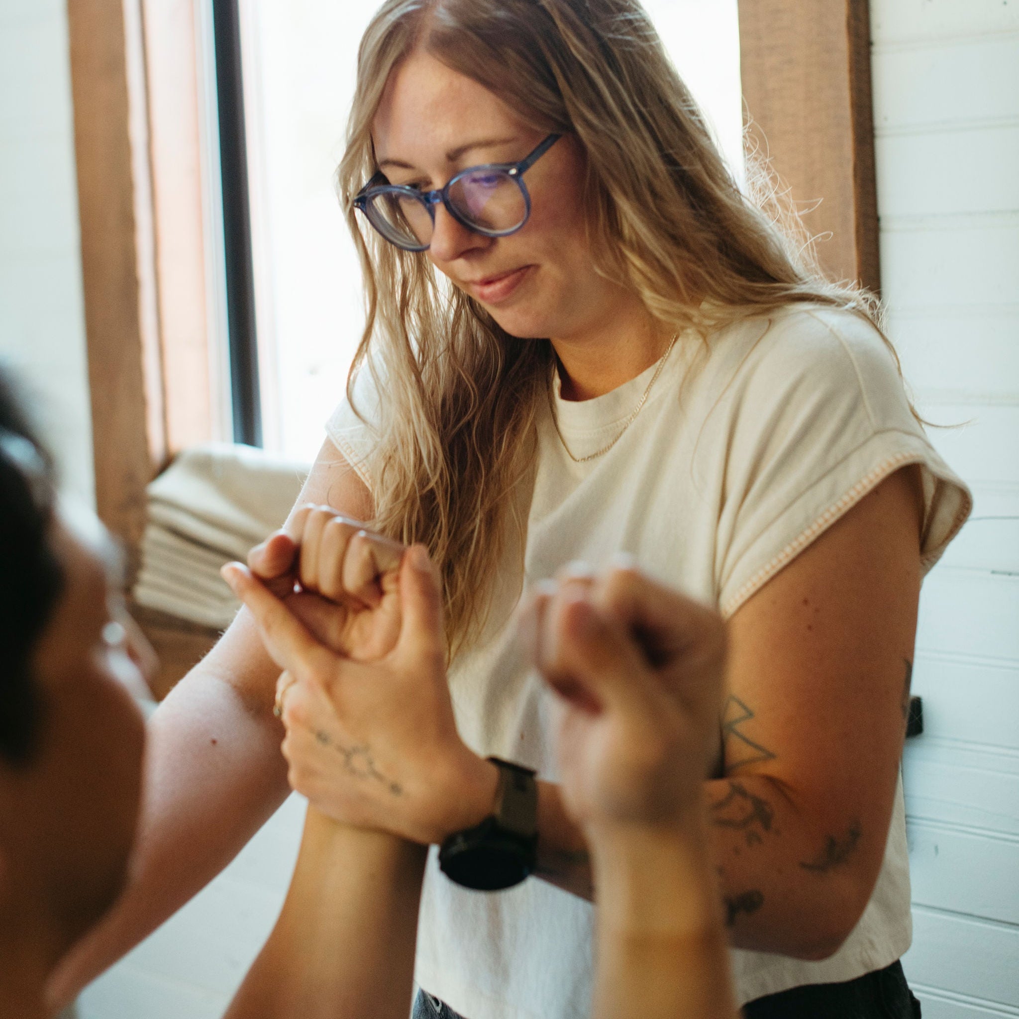 A woman with long blonde hair and blue-framed glasses wearing a white t-shirt and dark jeans, looking down while another person's hand is held by her. The scene is set in a brightly lit room with a white paneled wall and wooden trim.